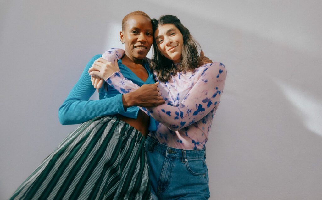 A Black woman with a blue long sleeve shirt and green skirt being hugged by a light-skinned woman with short hair wearing pink long sleeve shirt and blue jeans