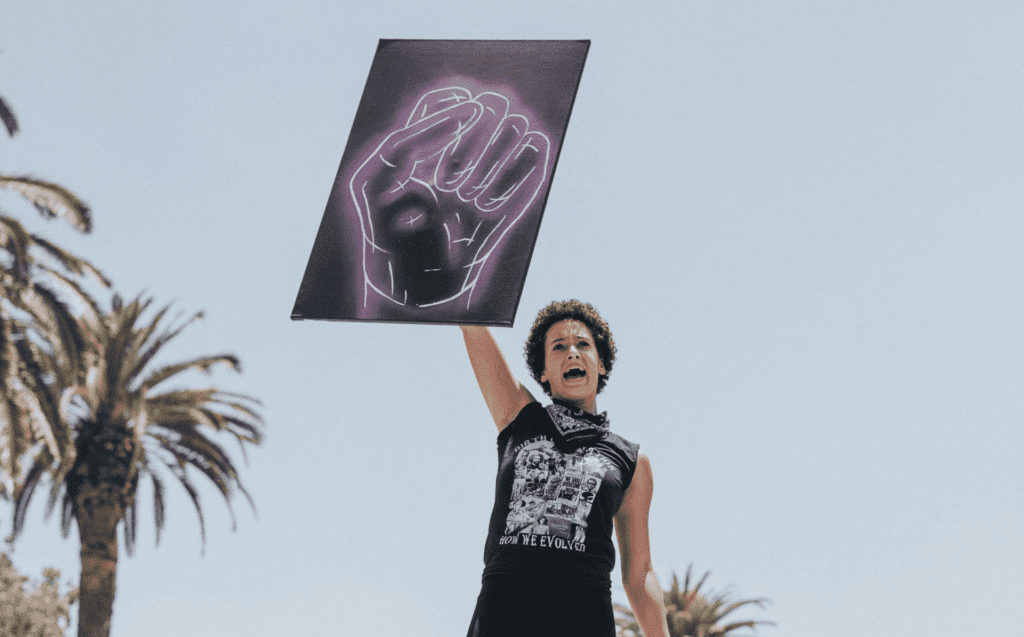 Black woman in the daytime on top of car with a poster of a fist during the Black Lives Matter protests. There are palm trees behind her.