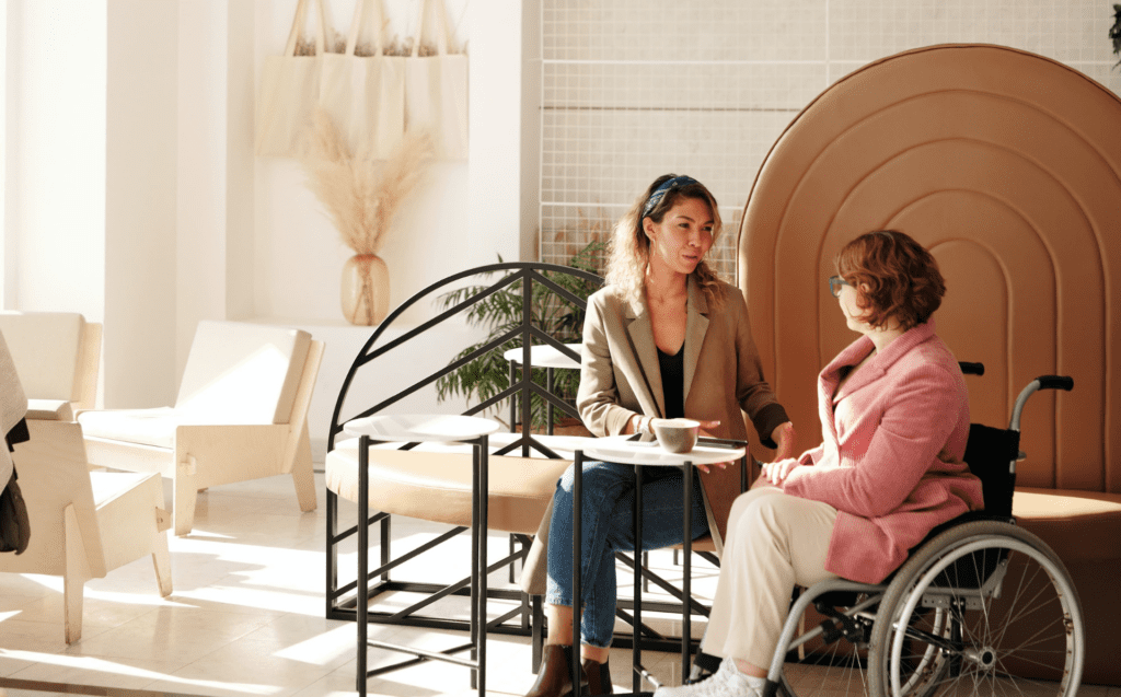 Two women talking at a table at a minimalist lounge. One woman is in a wheelchair, has short red hair, a pink jacket and white pants. The other woman is holding a cup of coffee and is wearing a great jacket with blue jeans. She has longer blonde hair.