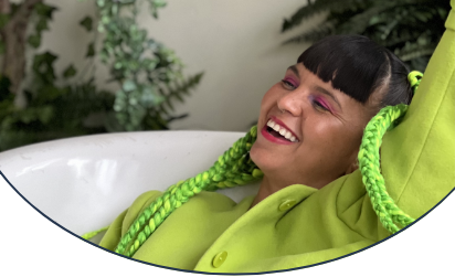 Li Saumet, singer of Bomba Estéreo, wearing a green jacket with green braids, laying a tub smiling surrounded by greenery