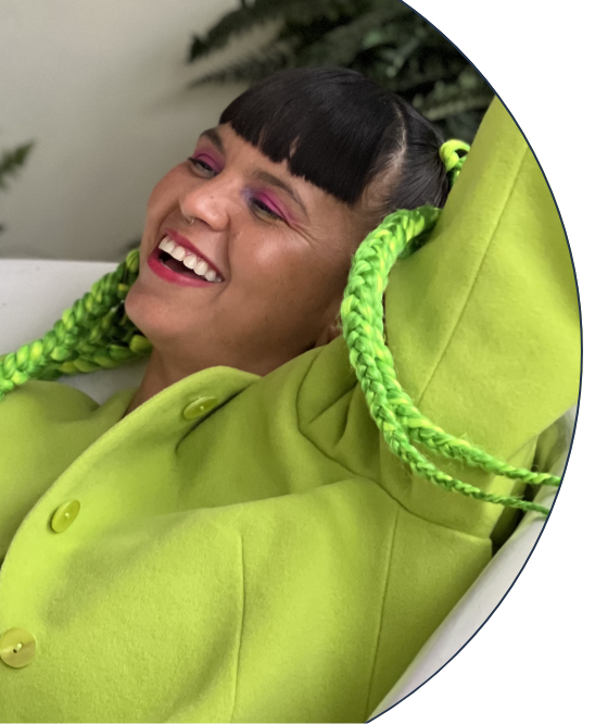 Li Saumet, singer of Bomba Estéreo, wearing a green jacket with green braids, laying a tub smiling surrounded by greenery