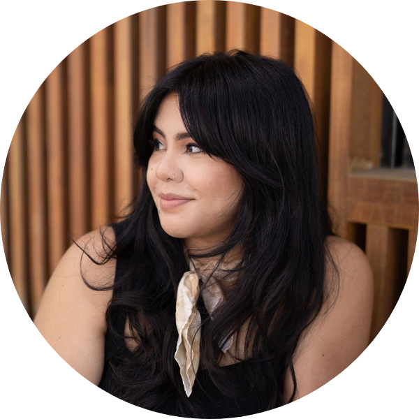 Priscilla Castro looking to the left as she sits in front of a brown panel wall. She has long black hair with curtain bangs, and is wearing a black tank top with a scarf on her neck. 