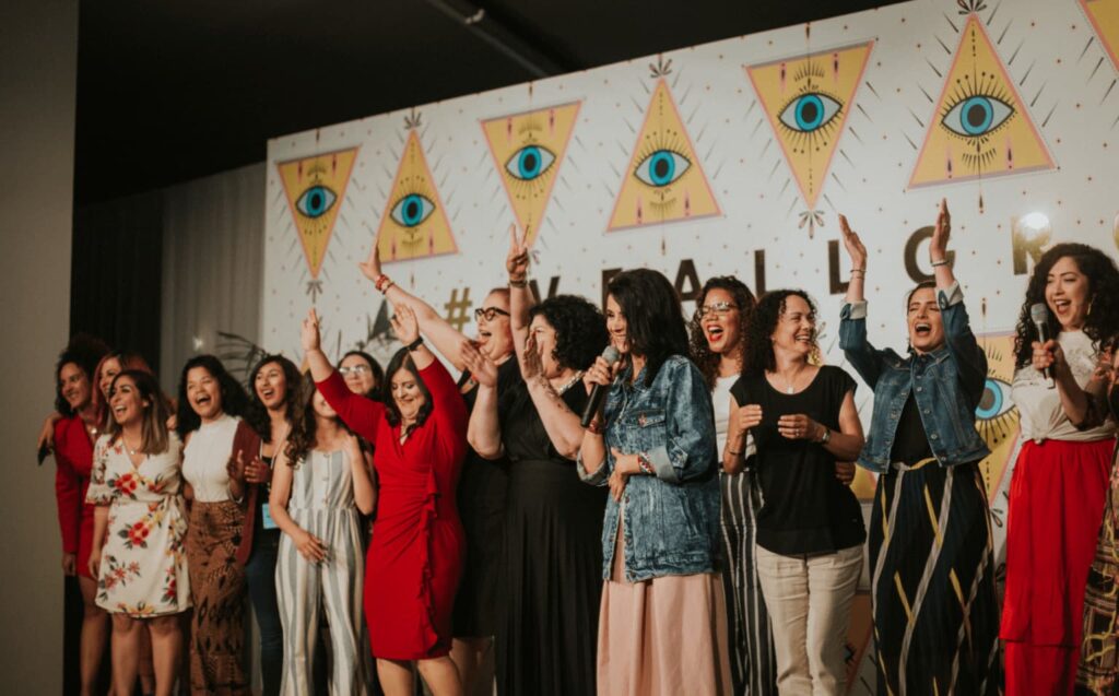 To show a perfect example of Amigahood, the photo shows a large crowd at the 2019 #WeAllGrow Summit after Storytellers, with all of them cheering, smiling and full of joy. Here, a lot of attendees joined Ana Flores on stage, where she is pictured in front of everyone wearing a denim jacket and a pink skirt, holding a microphone and talking to the audience.