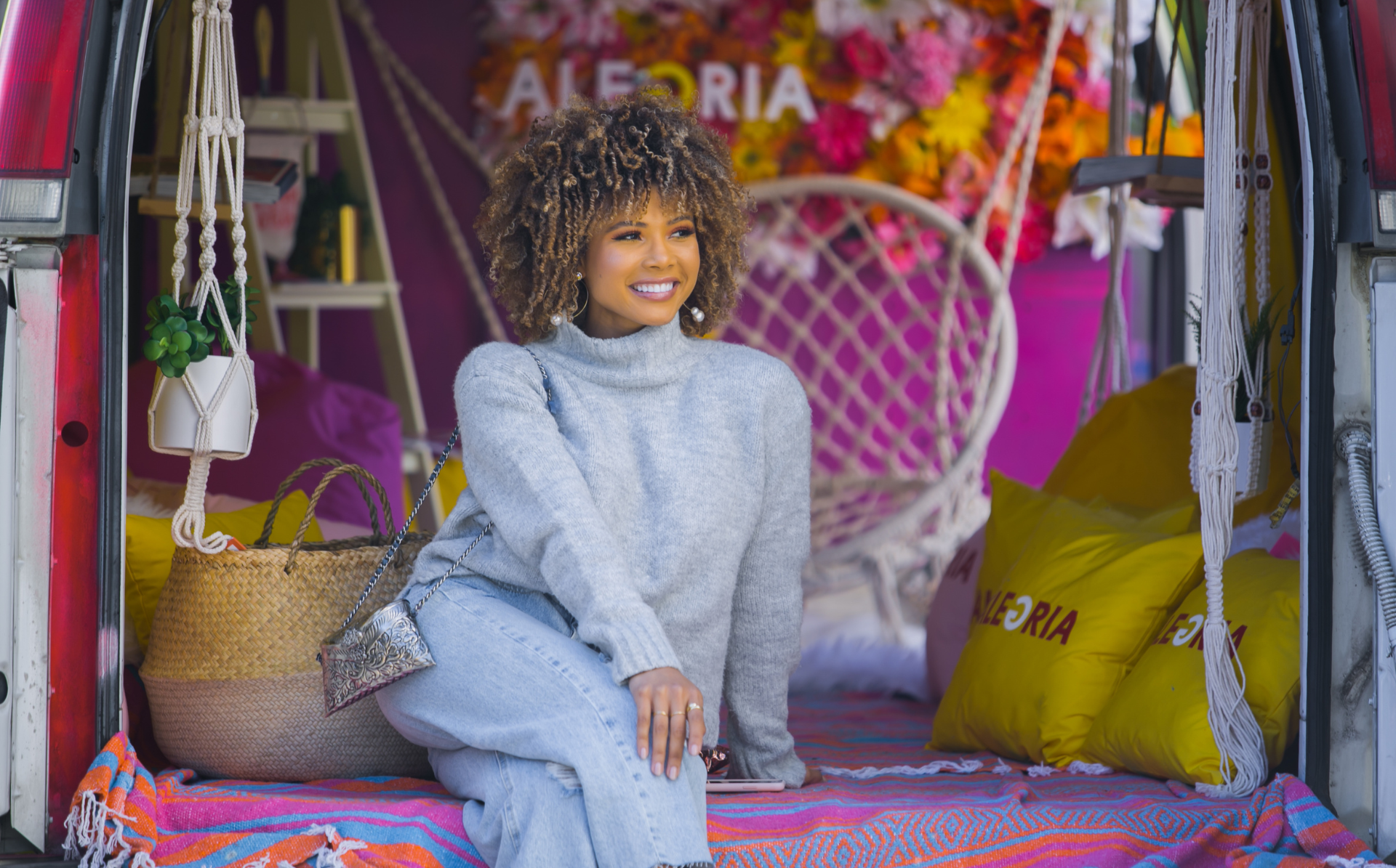 Pictured is a light skinned Afro-Latina woman sitting on an Alegria Publishing bus smiling away from the camera. She is wearing a grey turtleneck, and light denim pants. She has light brown and blonde hair in an afro.