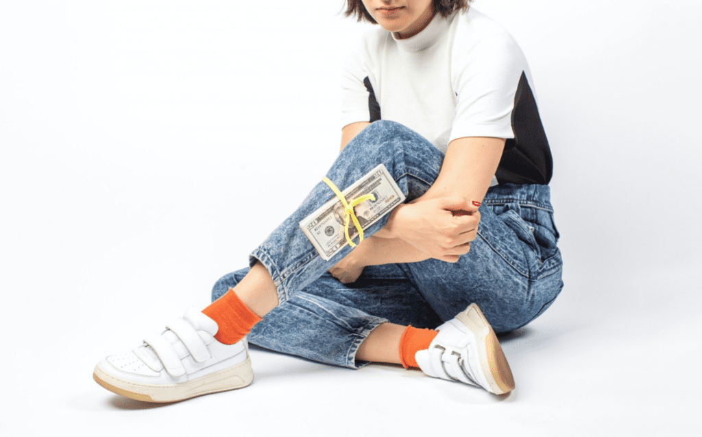 This is a photo of a girl sitting on the floor, with one foot out and a stack of money tied to her leg. She is wearing denim pants, a white and black shirt, has orange socks and white sneakers. Her face is cut off in the picture, but she has a short bob and you can see her lips.