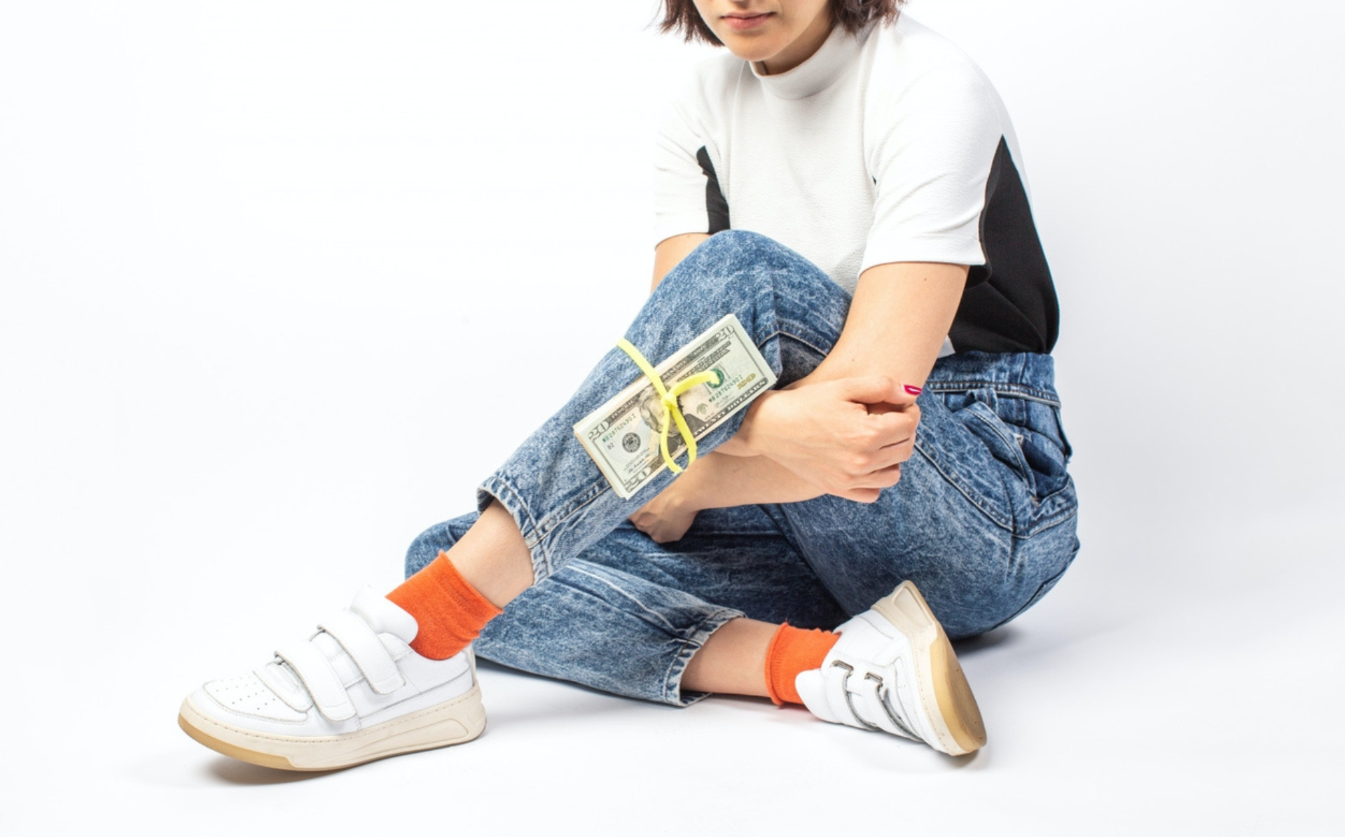 This is a photo of a girl sitting on the floor, with one foot out and a stack of money tied to her leg. She is wearing denim pants, a white and black shirt, has orange socks and white sneakers. Her face is cut off in the picture, but she has a short bob and you can see her lips.