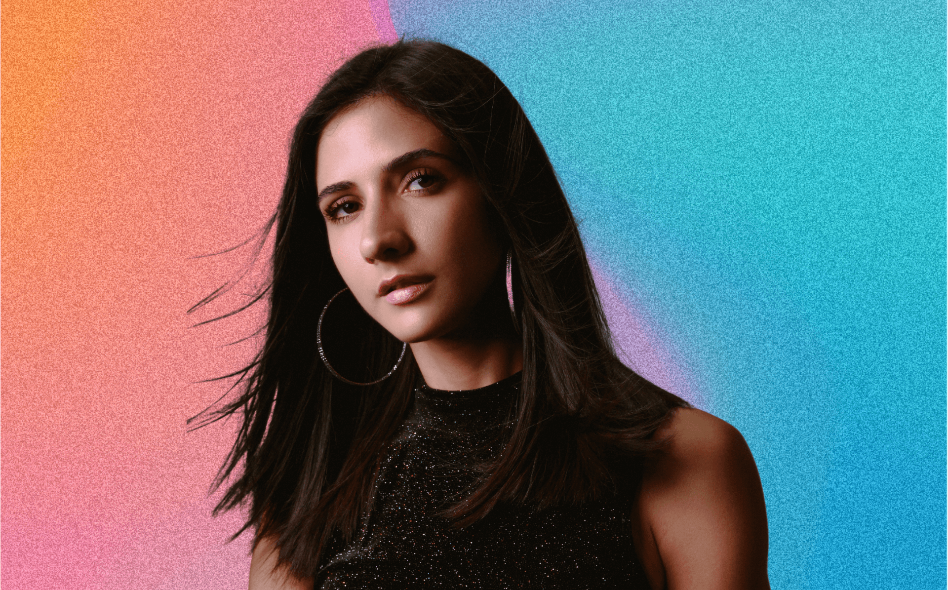 Disability activist and model Paula Carozzo is pictured in her headshot facing the camera with her hair being blown slightly by the wind. She is wearing large hops and a black shirt. Her picture is cutout to be in front of a pink and blue grainy gradient.