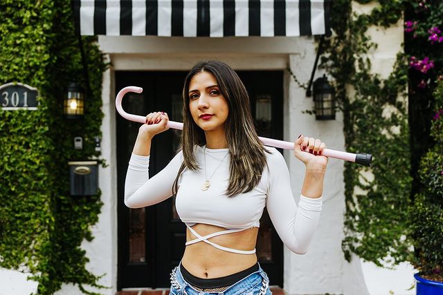 Disability activist and model Paula Carozzo is pictured in front of the entrance of a building wrapped in green wines, with a black and white striped awning over the entrance. She is pictured standing with a white crop top that wraps on her stomach, and blue jeans. She is holding her cane with two hands over her shoulders. 