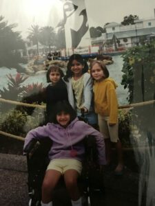 Paula is pictured as a child with her three friends behind her at Sea World in front of a sea world lighthouse that is surrounded by walruses. She is smiling with short hair at the camera sitting in a wheelchair in front of her friends, with a purple hoodie and lime green shorts. Her friend behind her at her left is with short hair and a black shirt, the one in the middle is holding the handle on Paula's wheelchair and she hair two pigtails, wearing a floral yellow shirt and a blue hoodie. Her friend on the right has short hair as well and is wearing a mustard yellow hoodie and cream colored capri pants. 