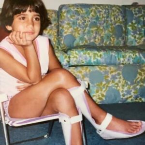 Paula is pictured as a child sitting on a foldable chair with her hand holding her head. She is crossing her legs, and she is wearing two leg braces. 