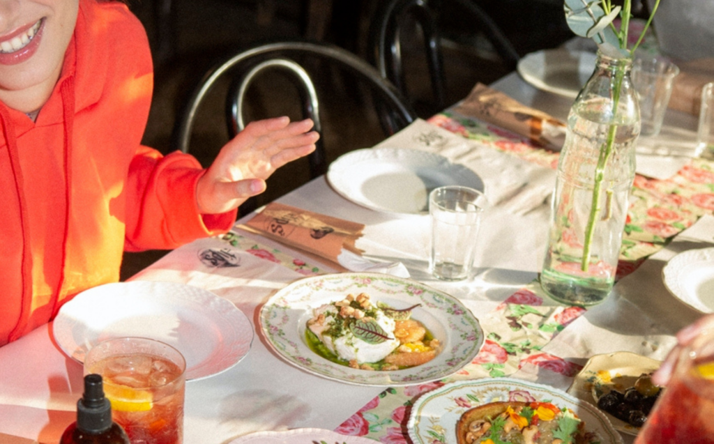 Woman smiling at the edge of a dinner table filled with plates, plant-based dishes and drinks. She is wearing a bright orange outfit during the daytime, and the sun is peeking through the room in what looks like golden hour.