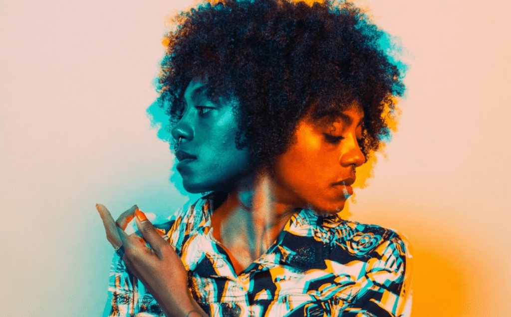 Image of woman with two faces, both faces looking in different directions to represent and personify imposter syndrome. The right face is in an orange tone and the left one is in a blue tone, and she is holding her hand up to her shoulder. She has curly short hair and has a black and white button up shirt.