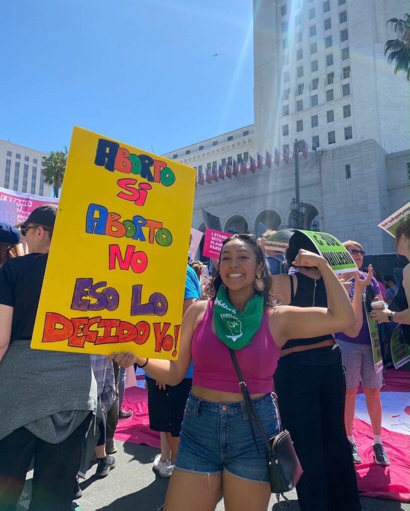 Maria Alejandra Paz at an abortion rally outside of the Los Angeles city hall with a poster the says, "Aborto is, aborto no, eso lo decido yo." She is wearing a fuschia halter top with denim shorts, and she is wearing a green bandana on her neck. She has her left arm up flexing her muscle, and is smiling at the camera.