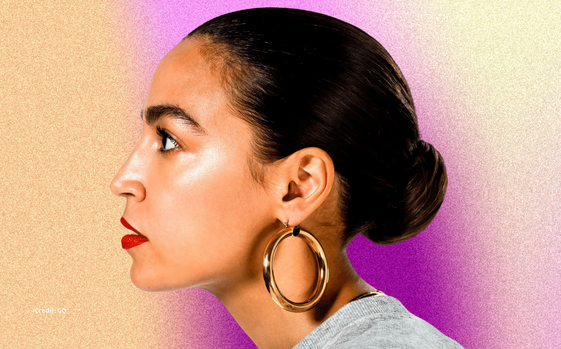 A side profile of Congresswoman Alexandria Ocasio-Cortez (D-NY-14), as shot by GQ. She is facing the left, has her hair in a slicked back low bun. She is wearing a red lip with a natural glow to her face, and is wearing large gold hoops. She is wearing a grey sweatshirt. Behind her is a peach and purple gradient grainy background.