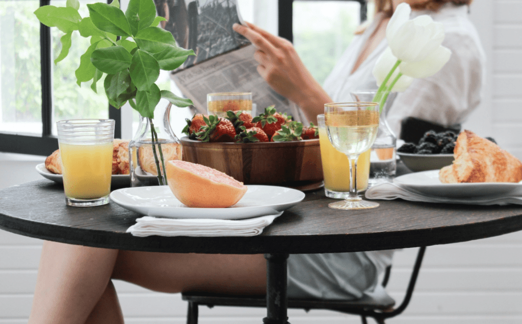 Woman having her morning routine sitting at her dining table reading the newspaper. The table has a bowl of strawberries, half a grapefruit on a plate, scones, cups with orange juice, and a glass vase with some leaves in it.