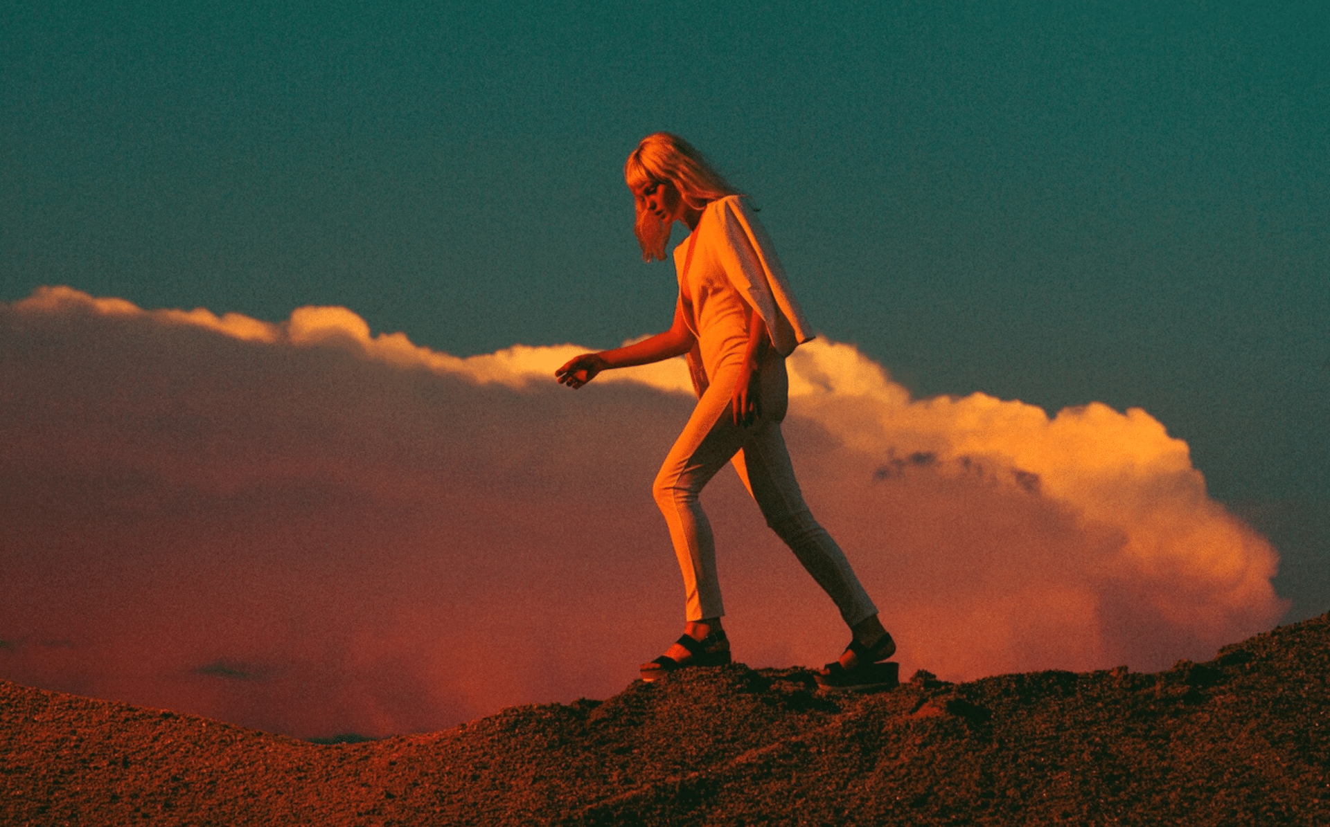 A woman with short blonde hair is hiking up a hill. She is wearing all white clothes, with a jacket draped over her shoulders. It is dusk and the golden hour light of the sun is hitting her. Behind her there is a massive red-tinted cloud with the blue sky peeking behind it.