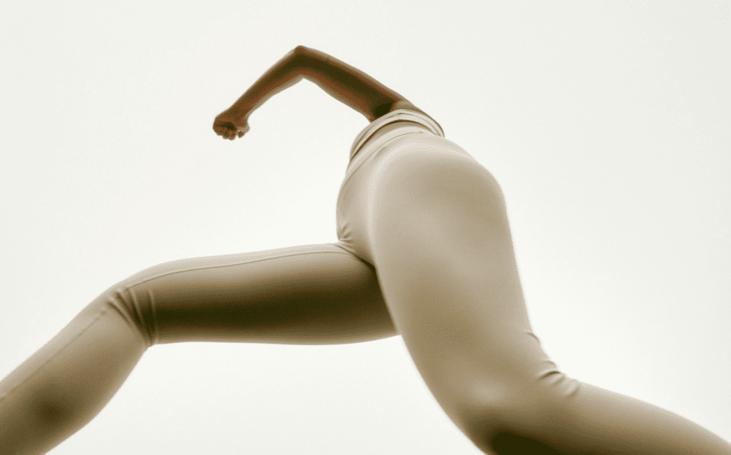 A black woman wearing ivory workout clothes takes an active leap over a camera.