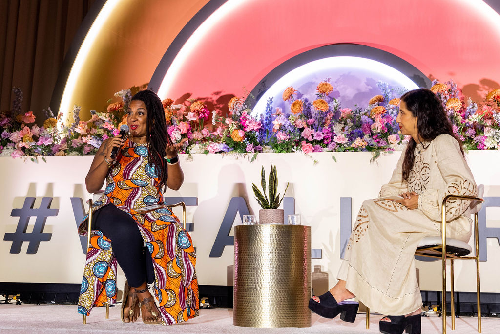 C-Suite leader and founder of Afro-Resistance Janvieve  Williams-Comrie and Editor in Chief of Vogue Mexico Karla de Salas sit on stage at #WeAllGrow Summit. Janvieve is wearing a long African print pattern dress with a high slit over a pair of black leggings. She has long dreads to the side of  her head and red lipstick on. She is speaking to the mic and she looks to the audience with her hand up. Karla Martinez de Salas looks at her, as she wears a long linen dress with pattern cutouts on the sleeves and down the center of the dress. She has long brown hair, and is wearing platform sandals. 