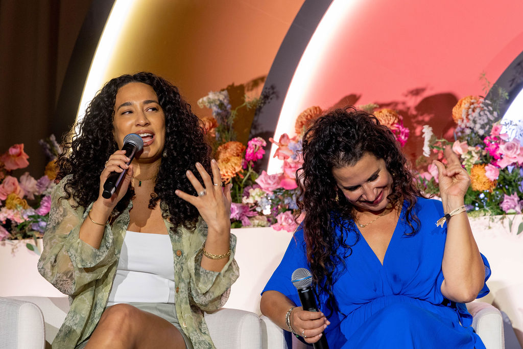 C-Suite leaders Julissa Prado (CEO and founder of Rizos Curls on the left and Susie Jaramillo (Founder of Encantos) on the right speaking on stage at #WeAllGrow Summit. Julissa Prado is wearing a green shirt with a white undershirt, and a matching green skirt. She has mid-length natural and perfectly coiled curls. She is holding a microphone and speaking into it. Susie Jaramillo is sitting to her left and is wearing a long blue dress, with long curly hair in a half up-do. She is snapping her fingers and smiling looking down in agreement with what Julissa is saying. 