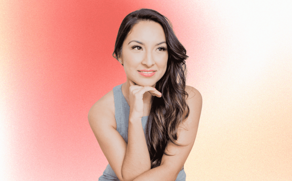 Business owner and sexologist Rebecca Alvarez Story is pictured in front a gradient peach and pink background, with her hand on her chin. She has long brown hair and is smiling away from the camera.