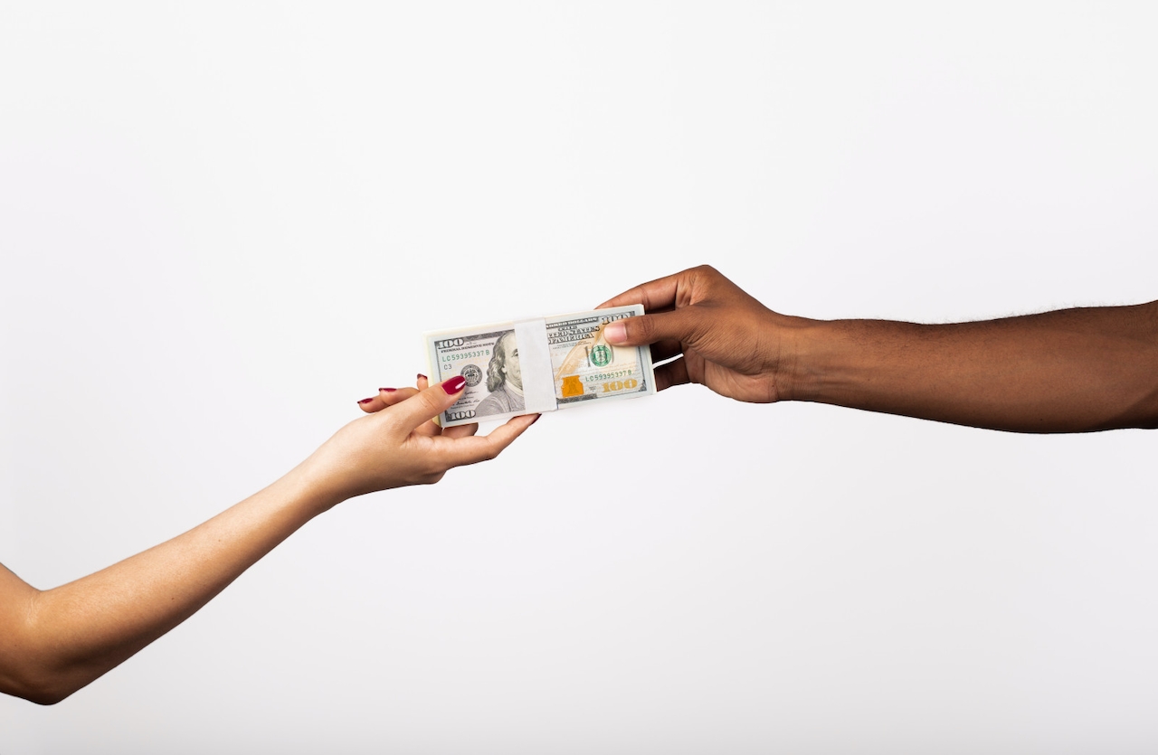 Two arms (one a light-skinned person and one a Black person) hold a stack of cash in between them. This illustrates the negotiation between brands and creators when negotiating brand deals.