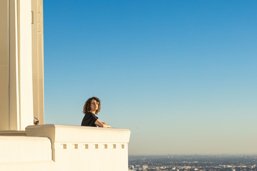 Gaby Moreno leans on the ledge of the Griffith Observatory, overlooking the skyline of Los Angeles, and faces the camera. 