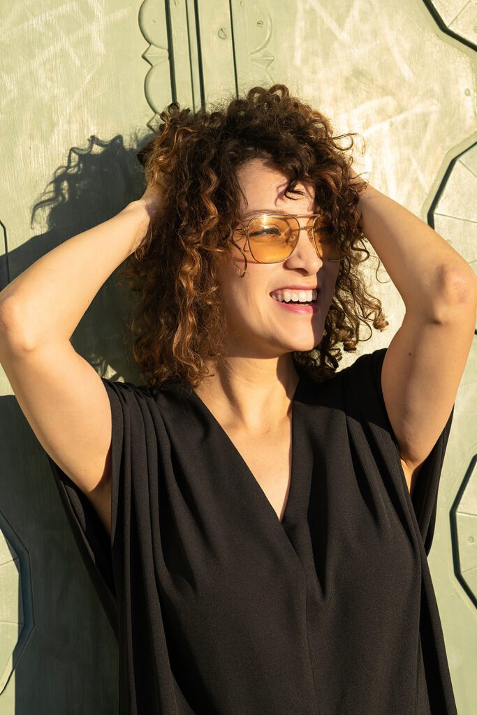 Gaby holds her hair as she smiles away from the camera as she is photographed in Griffith Park in Los Angeles, CA. She is wearing a black jumpsuit, with yellow tinted glasses, and she pictured behind a sage green door. 