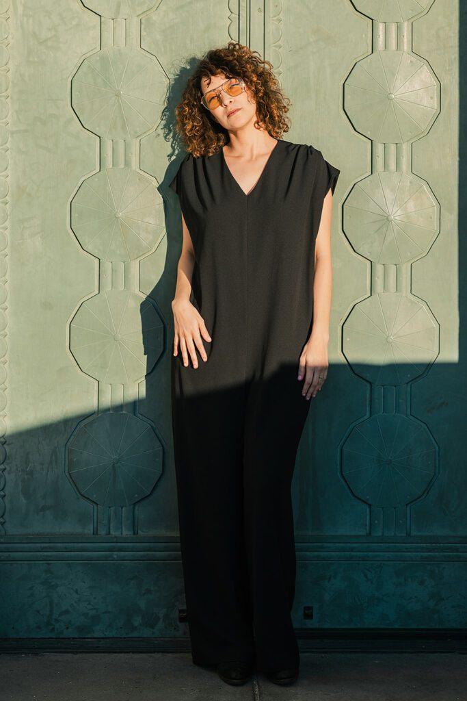 Gaby Moreno poses in front of a sage green door, as she is photographed in at the Griffith Observatory in Los Angeles, CA. She is wearing a black jumpsuit and with yellow tinted glasses.