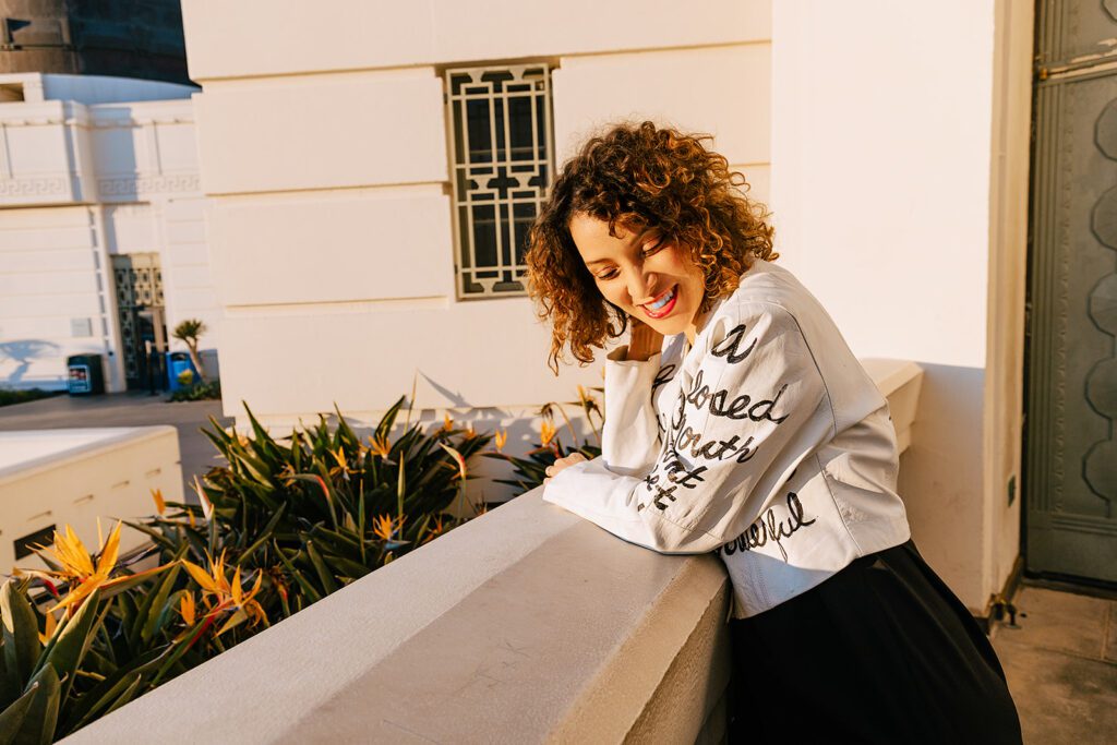 Gaby shyly smiles from the camera as she is photographed in Griffith Park in Los Angeles, CA. She is wearing a white leather jacket that is written on, and wears her natural brown and highlighted curls that are even more highlighted by the golden hour light. 