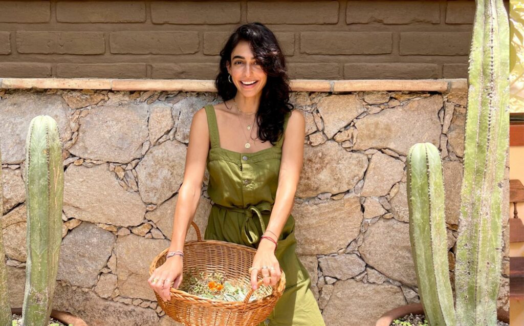 Creator of Water Thru Skin and slow living entrepreneur Valeria Hinojosa stands against a natural colored rock wall. She has mid-length hair, and is wearing a green jumpsuit with straps. She is holding a woven basket with leaves on it, and to her right and left are two cactus plants.