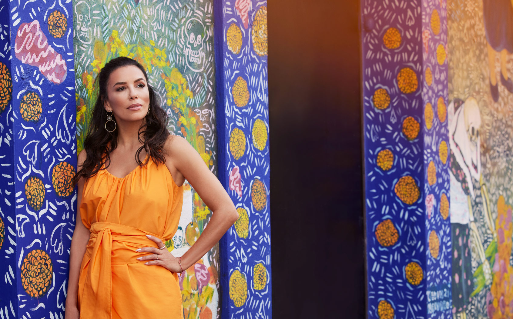Eva Longoria is pictured in Mexico filming for Searching For Mexico, wearing an orange dress, with her hair a little over her shoulders, and she has her hand on her hip. The background is a painted floral wall against a cobalt blue background. The flowers are marigolds painted in a pattern, with skulls scattered on the wall.
