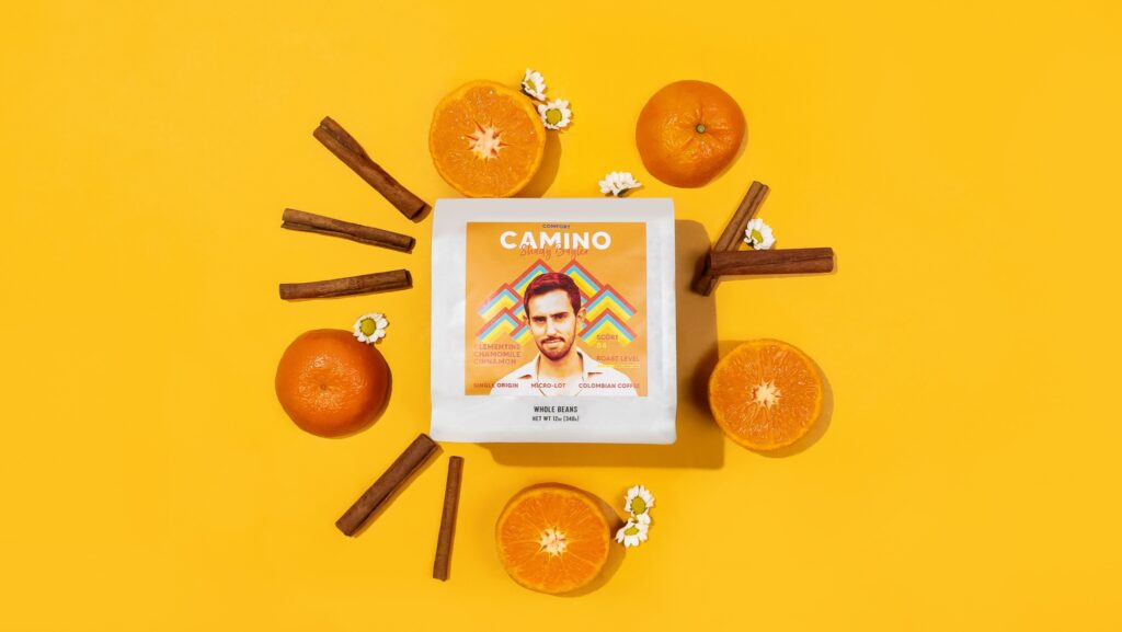 A package of Progeny Coffee is surrounded by oranges and cinnamon on a bright yellow backdrop. The coffee farmer is portrayed on the front of the Coffee package matching with the same yellow. 