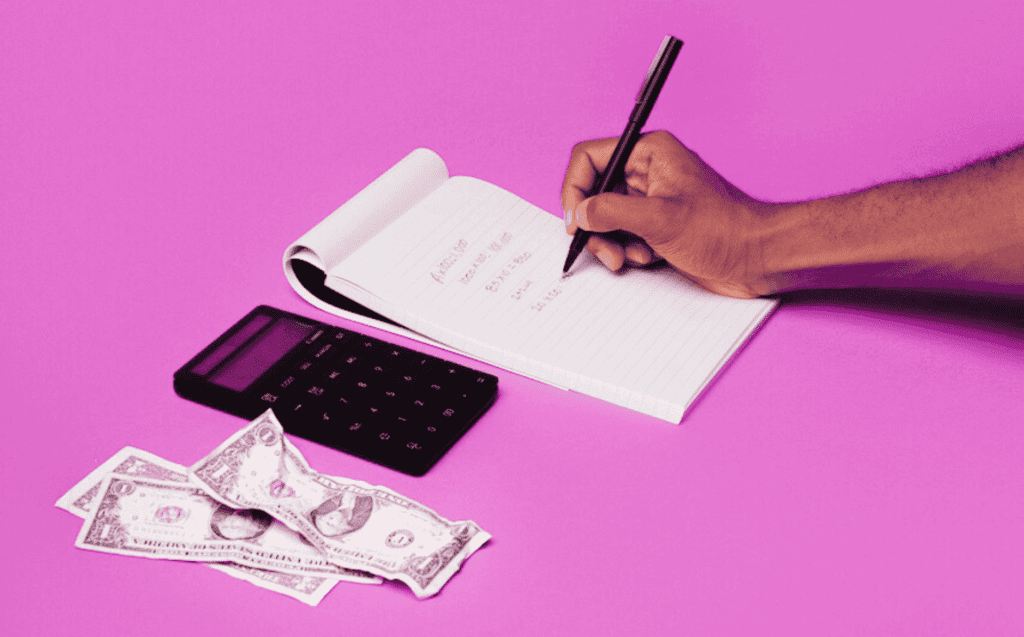 To illustrate the budgeting tips listed in this article put in action, a hand is seen writing on a note pad alongside a calculator and a few single dollar bills. The image is seen against a bright purple background.